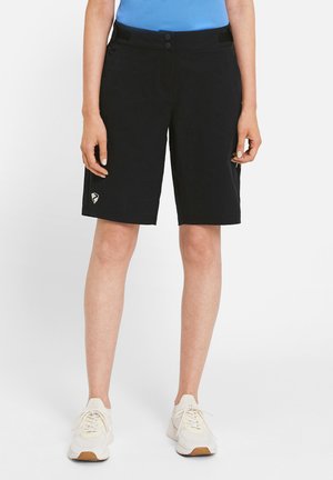 Person trägt schwarze knielange Shorts mit Logo am linken Bein, steht, kombiniert mit weißen Turnschuhen und blauem Oberteil.