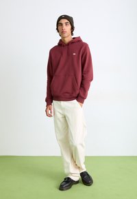 Sweat à capuche bordeaux avec poche avant et cordon de serrage, associé à un pantalon crème rayé et des chaussures noires épaisses. Le fond présente un sol vert.