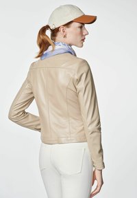 Beżowa skórzana kurtka o dopasowanym kroju, z widocznym szwem i gładką teksturą. Osoba nosi ją w zestawieniu z białymi spodniami i jasnoniebieskim szalikiem.