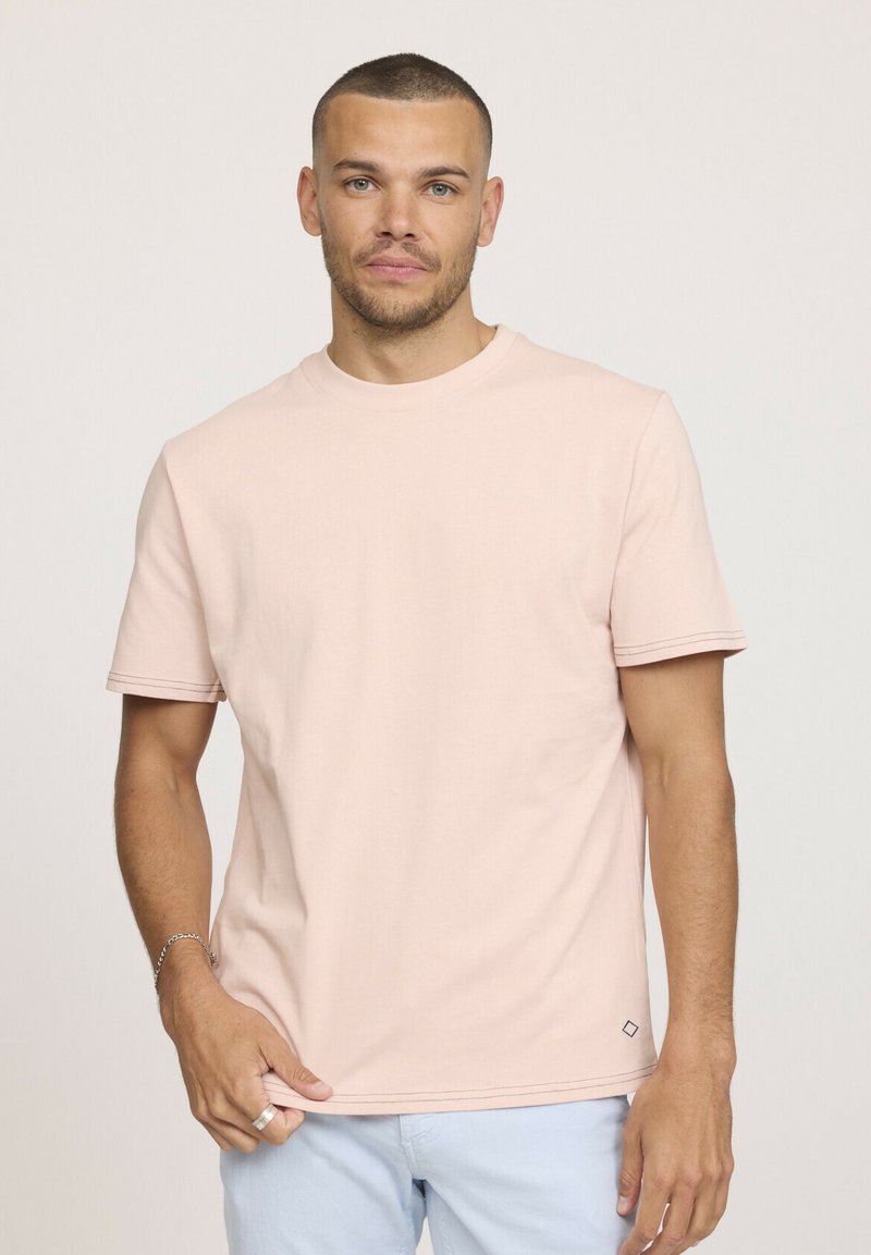T-shirt léger à manches courtes de couleur rose pâle en tissu doux ; présente un col rond et un ourlet double couture avec un détail de logo discret sur le côté.