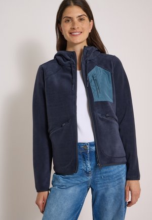 Kvinde iført en mørkeblå hættetrøje med lynlås og en turkis brystlomme, hvid skjorte og blå jeans, smiler til kameraet mod en ensfarvet baggrund.