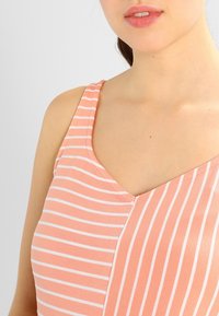 Nainen, joka pukeutunut hihattomaan persikansävyiseen toppiin, jossa vuorottelevat vaakasuorat ja pystysuorat valkoiset raidat, näkyvissä kaula-aukko ja osa kasvoista.