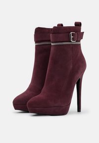 Pár burgundských semišových kotníkových bot s platformovými podpatky, dekorativní stříbrný zip kolem kotníku a zapínání na přezku.