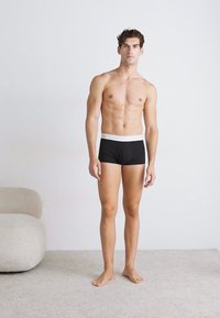 Zwarte boxers met een witte tailleband, aansluitend ontwerp en zachte textuur. Model staat blootsvoets tegen een effen achtergrond.