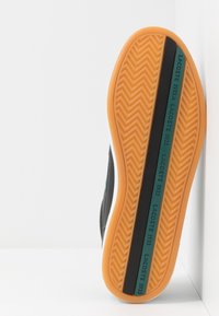 Suola di sneaker nera con motivo a spina di pesce, con base in gomma arancione e una striscia verde scuro con la scritta "LACOSTE 1933."