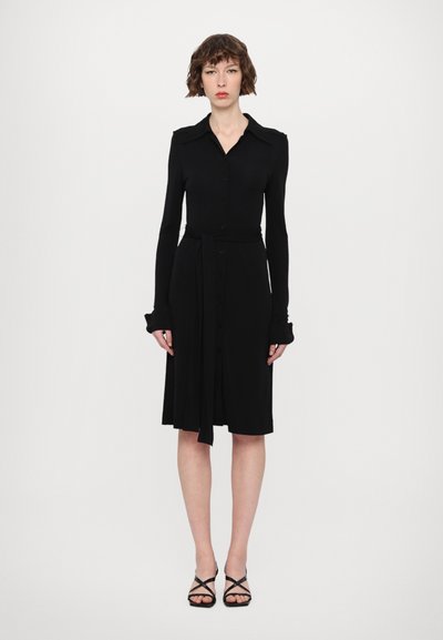 Čierne šaty s dlhým rukávom, prispôsobeným telom, zapínaním na gombíky vpredu a opaskom na viazanie. Majú špicatý golier a rozšírený lem s širokými manžetami.