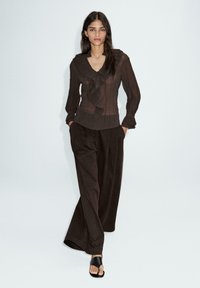 Blouse noire texturée avec un détail volants au niveau du col, associée à un pantalon large marron foncé ample et des sandales noires à enfiler.