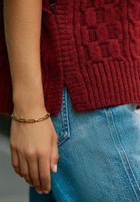 Pull en tricot bordeaux avec motifs texturés, associé à un jean bleu. Un bracelet chaîne en or avec des maillons rectangulaires est porté au poignet.