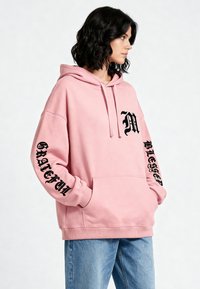Felpa rosa con maniche lunghe, tasca frontale, cappuccio con cordino e dettagli in testo nero sul davanti e sulle maniche. Tessuto morbido; vestibilità oversize.