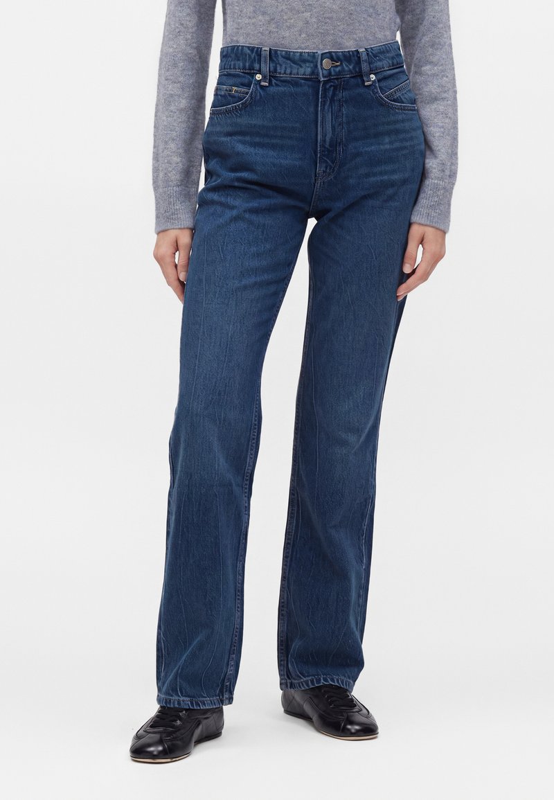 Personne portant un jean bleu foncé taille haute à jambes droites, un pull gris à manches longues et des chaussures noires à lacets sur fond uni.