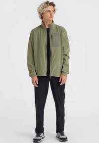 Green fleece jacket with a high collar, front zip, and chest pocket. Black pants and gray athletic shoes complete the outfit.