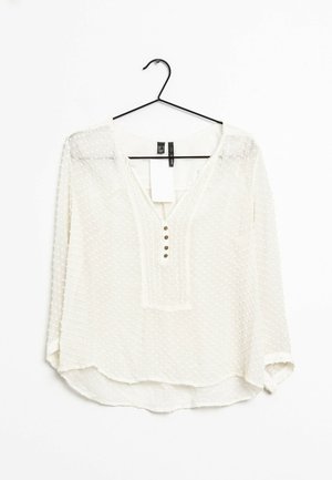 Blusa blanca con puntos de polka texturizados, escote en V y tapeta de tres botones. Mangas largas con puños ligeramente doblados. Tejido ligero y transparente.