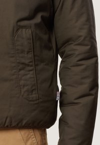 Oliven grøn polstret jakke med en lynlås-lomme, der har en tekstureret overflade og kontrasterende syninger. Logoet er synligt på ærmet.