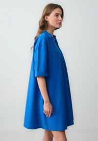 Abito a vestito morbido di un blu vivace, realizzato in tessuto liscio. Presenta un colletto e maniche corte arrotolate, con un orlo curvo.
