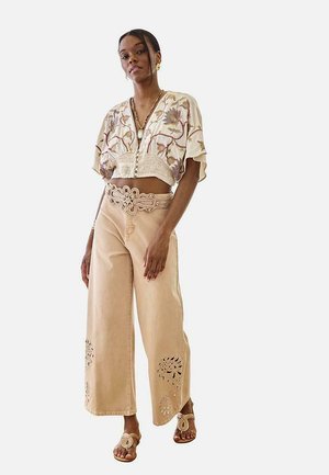 Mujer con pantalones anchos beige con patrones recortados, blusa corta floral, cinturón decorativo y sandalias a juego, de pie sobre fondo blanco.