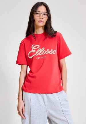 Femme aux longs cheveux noirs portant des lunettes, un t-shirt rouge "Ellesse EST 1959" et un pantalon de survêtement gris clair sur un fond clair uni.
