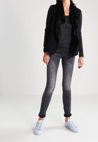 Kobieta nosząca czarną otwartą kurtkę z puszystym kołnierzem, ciemnoszare dopasowane dżinsy oraz białe sneakersy, stojąca na jednolitym tle.