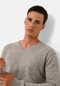 Grauer V-Ausschnitt-Pullover aus weichem Strickstoff mit gerippten Bündchen, der eine leichte Textur und eine lässige Passform aufweist. Das Modell posiert mit den Händen nahe den Ärmeln.