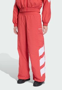 Červené atletické nohavice s bielymi diagonálnymi pruhmi po stranách, vyrobené z ľahkého, štruktúrovaného materiálu, s elastickým pásom.