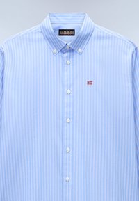 Chemise à manches longues bleu clair avec des rayures blanches, col boutonné et petit logo brodé. Le tissu a une texture lisse et une coupe classique.