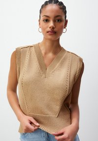 Noor naine punutud juustega kannab kuldseid rõngaste kõrvarõngaid ja beeži ilma varrukateta kudumisvesti helesiniste teksade peal.