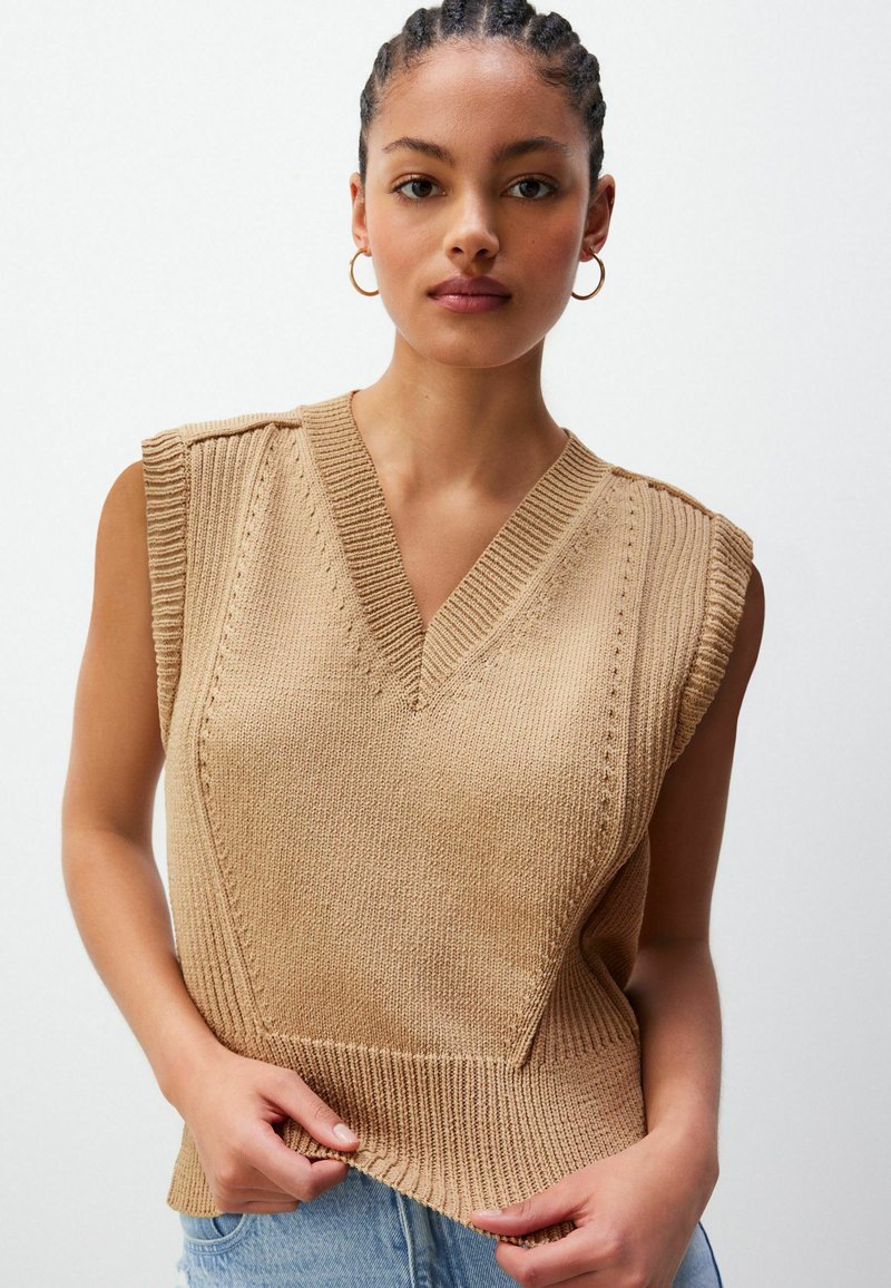 Noor naine punutud juustega kannab kuldseid rõngaste kõrvarõngaid ja beeži ilma varrukateta kudumisvesti helesiniste teksade peal.