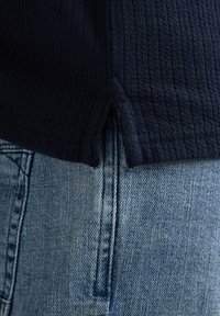 Haut en tissu côtelé bleu marine foncé avec fente latérale, associé à un jean en denim bleu clair présentant une couture de poche visible et un tissage texturé.