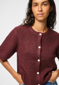 Bordeaux gebreide cardigan met korte mouwen, voorzien van een ronde halslijn en vijf witte knopen aan de voorkant. Textuur van de stof is duidelijk zichtbaar.