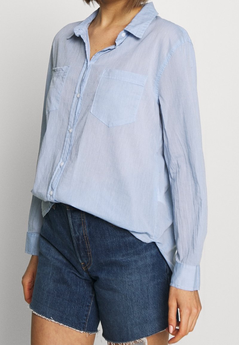 Femme portant une chemise bleu clair à manches longues boutonnée et un short en jean bleu foncé avec des bords effilochés sur un fond uni.