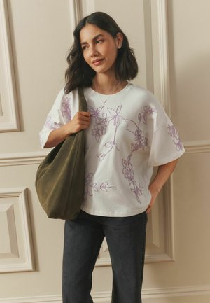 Woman with shoulder-length dark hair wearing a white floral embroidered shirt, black jeans, and holding a large olive green bag.