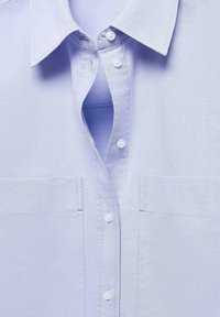 Light blue button-up shirt with a pointed collar, two front pockets, and a smooth, textured fabric. Features white buttons along the placket.