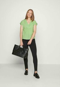 Top verde de mangas curtas com decote em V, combinado com leggings pretas. O modelo segura uma bolsa preta acolchoada e usa mocassins pretos.