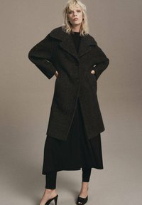 Abrigo de lana de cuadros en tonos oscuros con un gran cuello, cierre de botones y mangas oversized. Llevado sobre un vestido negro y leggings, con tacones.