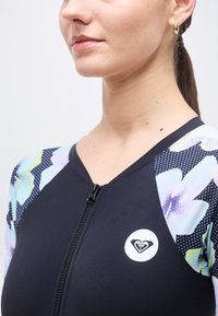 Γυναίκα που φοράει μαύρο τοπ με φερμουάρ και μανίκια με λουλουδάτο σχέδιο, με κυκλικό λογότυπο στο στήθος, με τα μαλλιά πιασμένα πίσω και χρυσό σκουλαρίκι κρίκο.