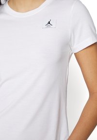 Hvid bomulds t-shirt med korte ærmer, prydet med et sort logo-patch på venstre bryst med en silhuet og signaturdetalje.