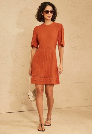 Vrouw draagt een oranje, gestructureerde jurk met korte mouwen, een zonnebril, houdt een kleine geweven handtas vast en draagt zwarte sandalen met gekleurde kraaltjes.