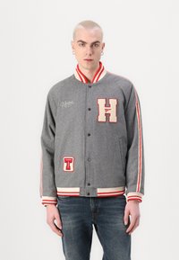 Szary wełniany kurtka typu varsity z czerwono-białymi paseczkami na kołnierzyku i ściągaczach. Zawiera duże haftowane naszywki "H" oraz "TOMMY". Zapinana na guziki z przodu.