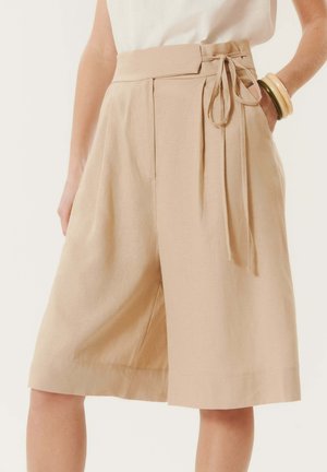 Short beige taille haute à jambes larges arrivant au genou, avec plis devant et ceinture à nouer sur le côté, porté avec un top léger et des bracelets.