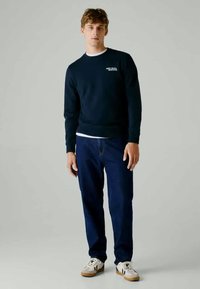 Sweatshirt azul-marinho com decote careca e logótipo branco, combinado com jeans azuis e sapatilhas brancas com detalhes pretos.