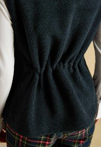 Schwarze Fleeceweste mit gerafftem Detail an der Taille, getragen über einem weißen Langarmshirt, kombiniert mit karierten Hosen.