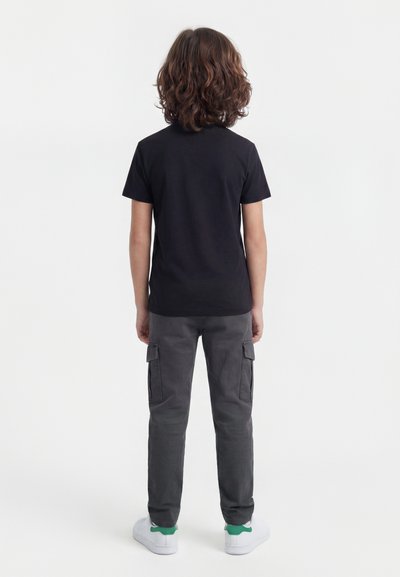 Παιδί με κυματιστά καστανά μαλλιά στέκεται με την πλάτη απέναντι, φορώντας απλό μαύρο μπλουζάκι, γκρι παντελόνι cargo και λευκά αθλητικά παπούτσια με πράσινες λεπτομέρειες.