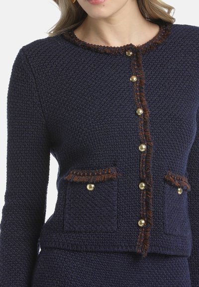 Navy textured cardigan with gold buttons, brown fringed accents on the collar and pockets, and two patch pockets at the chest.