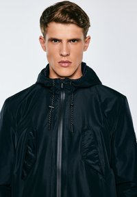 Jauns vīrietis ar īsiem gaiši brūniem matiem, valkājot melnu kapuci ar rāvējslēdzēju un auklām, baltā fona priekšā.