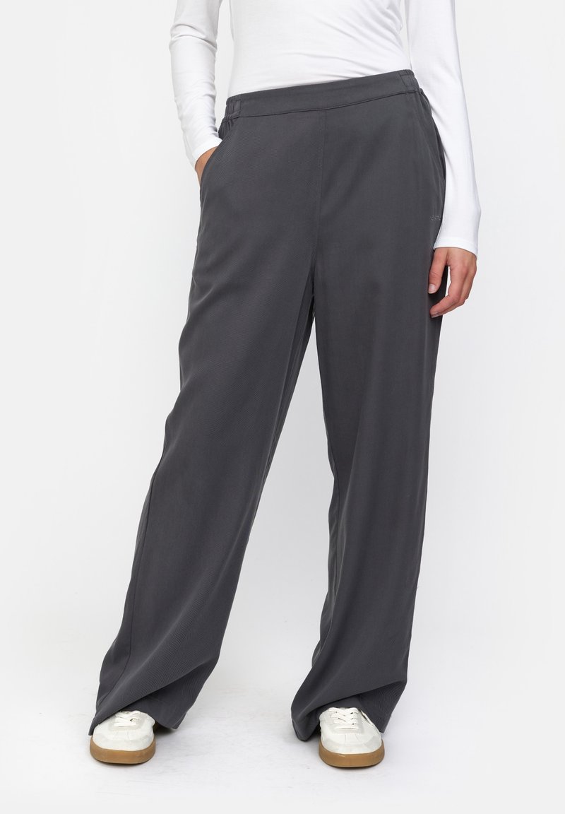 Personne portant un pantalon ample gris foncé, des baskets blanches et un haut blanc à manches longues, debout avec une main dans la poche.