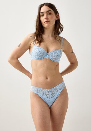 Kvinde med brunt hår iført matchende lyseblå blonder-bh og trusser, står med hænderne i siden mod en ensfarvet baggrund.