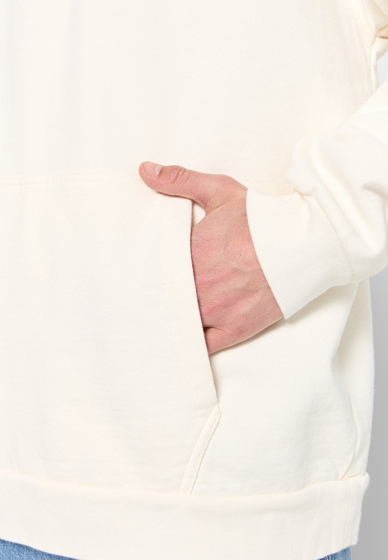 Helle beige Kapuzenjacke mit einer Kängurutasche, gerippten Bündchen und Saum. Der Stoff wirkt weich, die Strickstruktur ist sichtbar. Hand im Taschen.