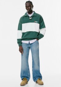 Pullover di pile a righe verdi e bianche con colletto, indossato sopra una camicia a righe rosa. Abbinato a jeans a gamba larga di un blu chiaro e stivali color sabbia.