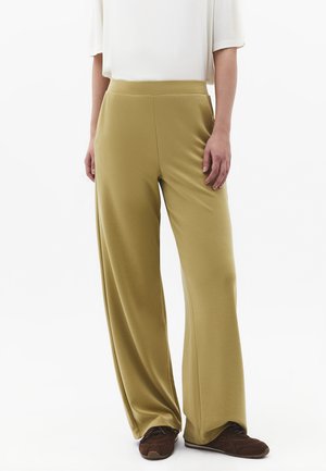 Femme portant un pantalon large olive, un haut blanc à manches courtes et des baskets marron, debout devant un fond blanc.