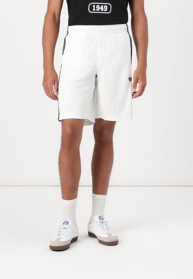 Shorts de sport blancs avec des rayures noires sur les côtés, en tissu texturé. Associés à un t-shirt noir et des chaussettes blanches avec des chaussures de couleur claire.
