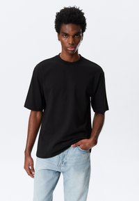 Mann iført en enkel svart oversized T-skjorte med lyseblå jeans, stående med en hånd i lommen mot en hvit bakgrunn.